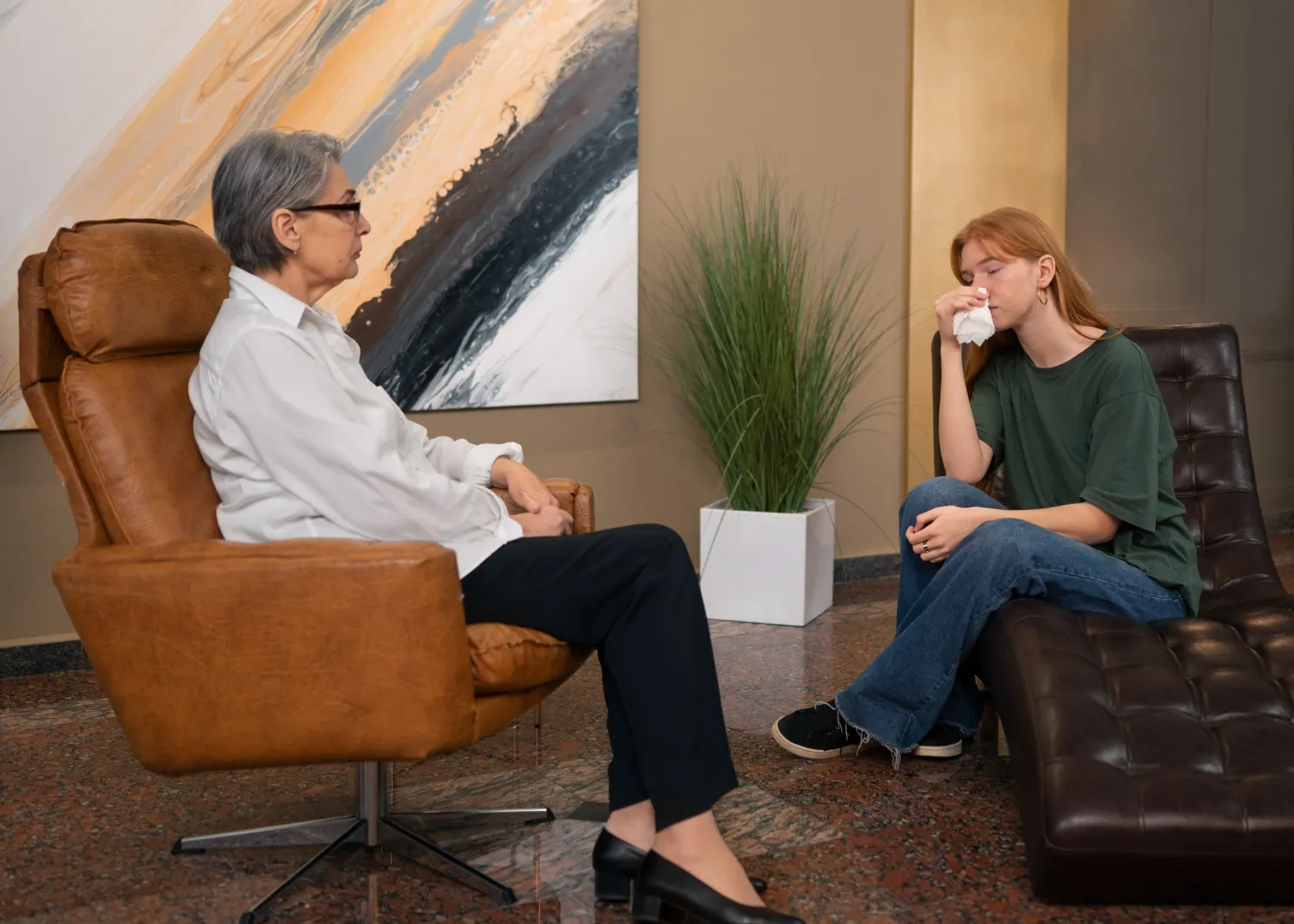 Sesión de terapia psicológica entre una mujer mayor y una joven, donde la joven muestra emociones mientras se limpia la cara con un pañuelo, en un ambiente acogedor con muebles modernos y una obra de arte abstracto en la pared.